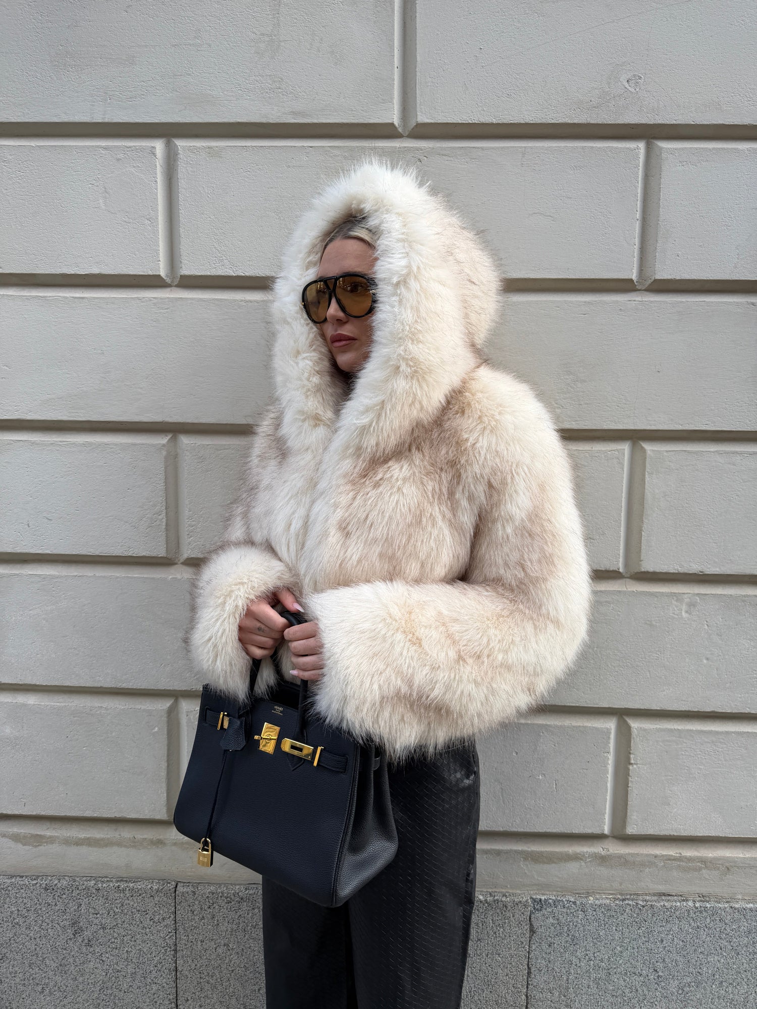 Person wearing a fur coat and sunglasses, leaning against a tiled wall.